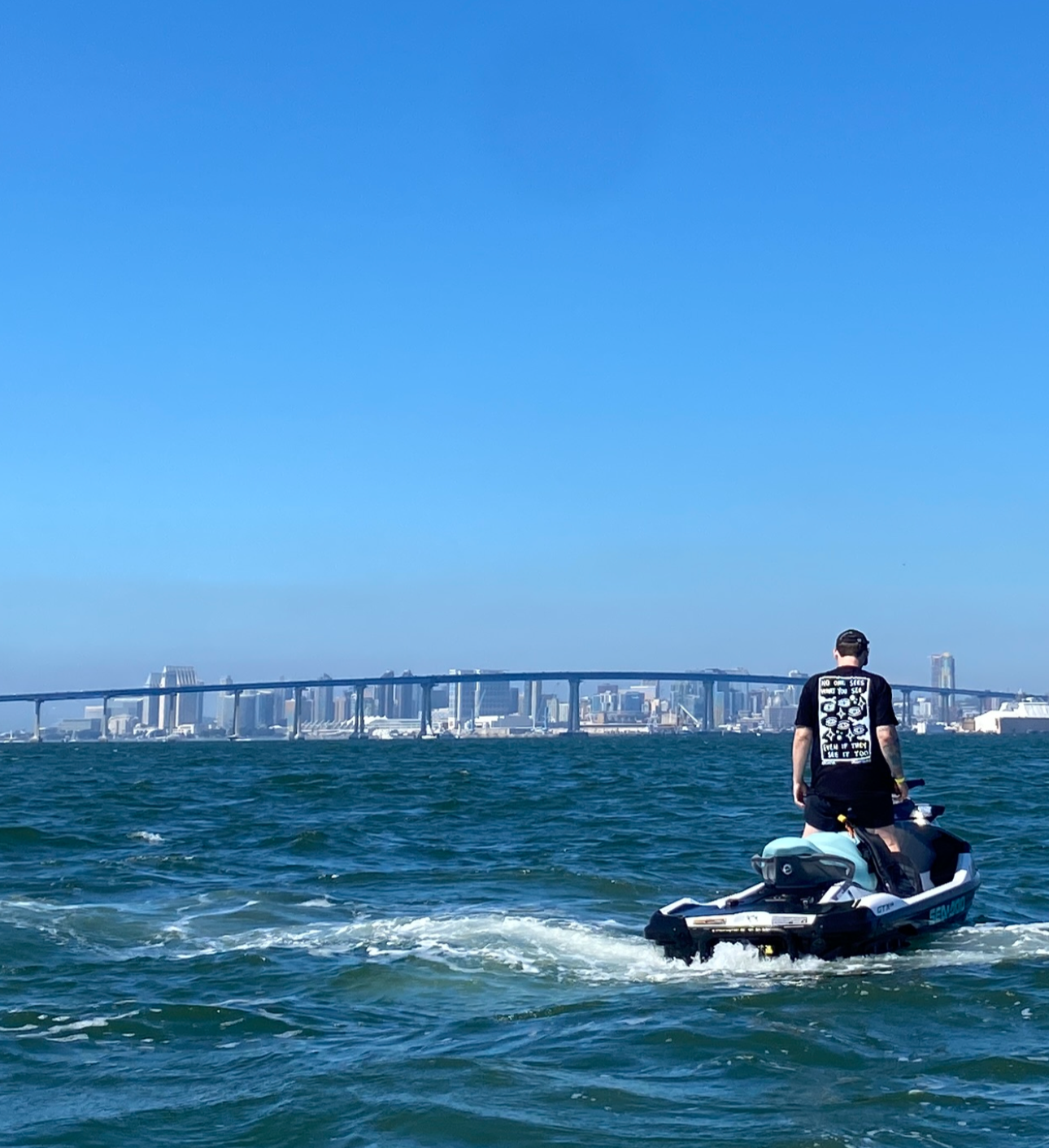 Person wearing the Skin Deep Vision Tee on a jet ski in the ocean with a clear blue sky and a bridge in the background spanning across the city of San Diego, CA.
